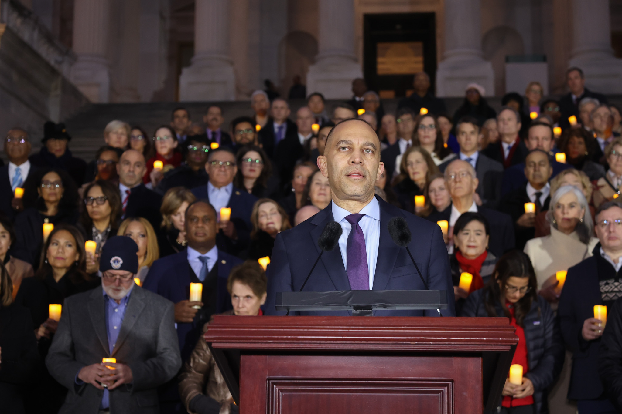 Leader Jeffries speaking from a podium in front of Members of Congress holding candles on the Senate steps