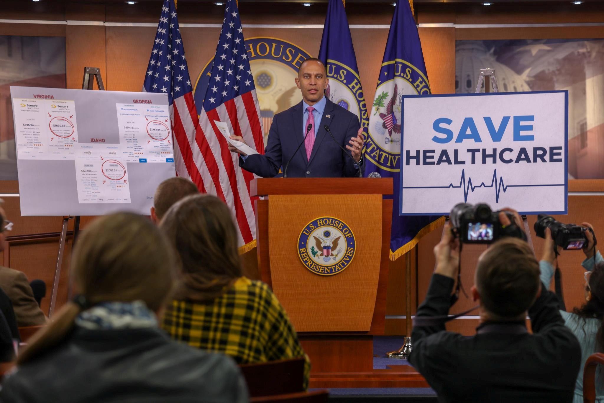 Leader Jeffries Speaking at Weekly Press Conference in between a sign that reads "Save Healthcare" and receipts showing increases in healthcare premiums
