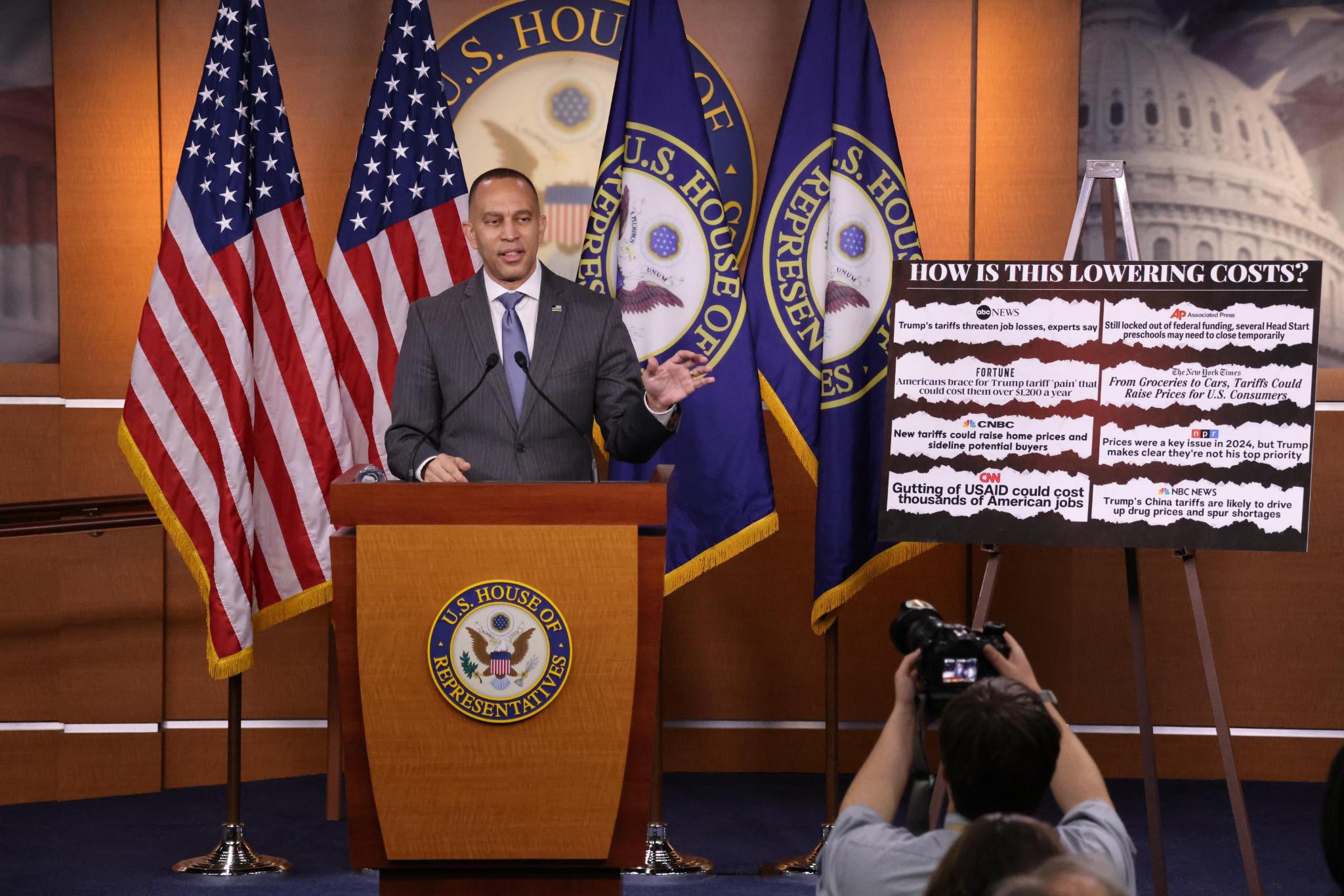 Leader Jeffries Speaking at Weekly Press Conference with a poster with the words "How is this lowering costs?" and headlines of far-right extremism