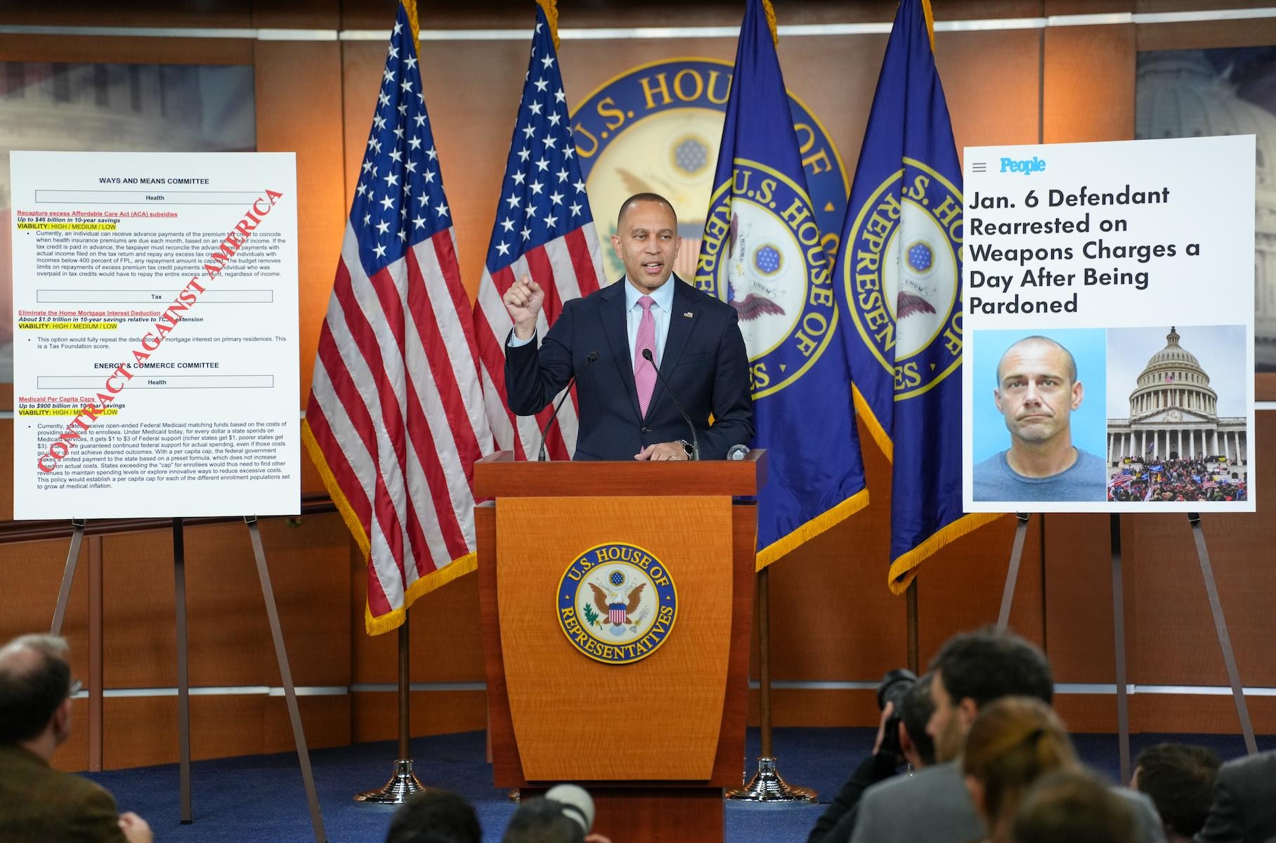 Leader Jeffries next to a poster with the words "Contract Against America" and a poster with a People Magazine headline: "Jan. 6 Defendant Rearrested on Weapons Charges a Day After Being Pardoned"