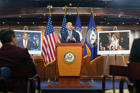 Leader Jeffries Speaking at Weekly Press Conference flanked by posters bearing images of Representative George Santos flashing peace signs (left) and Representative Marjorie Taylor Greene booing during the State of the Union